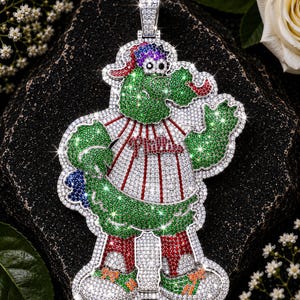 Könnte beinhalten: Ein silberfarbener Anhänger in Form einer Maskottchenfigur, die eine Baseballuniform mit dem Schriftzug Phillies auf der Brust trägt. Die Figur ist mit zahlreichen kleinen, funkelnden grünen, roten, blauen und klaren Steinen besetzt.