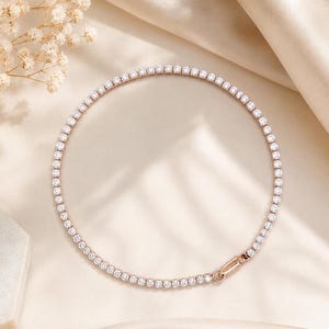 Könnte beinhalten: Eine Tennis-Halskette aus Roségold mit einer durchgehenden Reihe kleiner, runder, klarer und facettierter Steine in einer Metallfassung. Die Kette ist kreisförmig angeordnet und verfügt über einen sicheren, länglichen Metallverschluss am Ende.