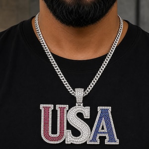 May include: A silver-toned curb chain necklace featuring a large pendant shaped as the letters USA. The U is encrusted with red stones, the S with clear stones, and the A with blue stones. The entire pendant and chain are covered in clear stones.