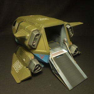 Puede incluir: Modelo detallado de un veh&iacute;culo de transporte militar en verde oliva, azul y gris. El modelo tiene el n&uacute;mero "002" impreso en el lateral y una rampa bajada. El veh&iacute;culo tiene un dise&ntilde;o cuadrado con m&uacute;ltiples motores y alas.