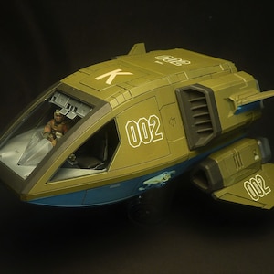 Puede incluir: Maqueta de nave espacial verde oliva y azul, con el n&uacute;mero "002" en blanco en el lateral y la cola. La cabina tiene una cubierta transparente y una figura es visible en el interior. La nave tiene un dise&ntilde;o futurista.