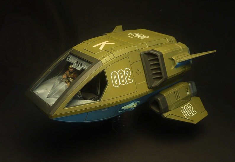 Puede incluir: Maqueta de nave espacial verde oliva y azul, con el n&uacute;mero "002" en blanco en el lateral y la cola. La cabina tiene una cubierta transparente y una figura es visible en el interior. La nave tiene un dise&ntilde;o futurista.