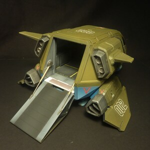 Puede incluir: Modelo detallado de un veh&iacute;culo de transporte futurista, principalmente verde oliva con detalles en azul y gris. El veh&iacute;culo tiene una rampa abierta, revelando un interior. El n&uacute;mero "002" es visible en el lateral.