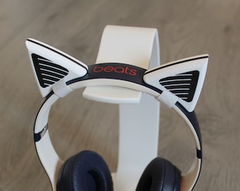 May include: White and blue cat ear headphones with a red "beats" logo on the headband. The headphones are on a white stand.