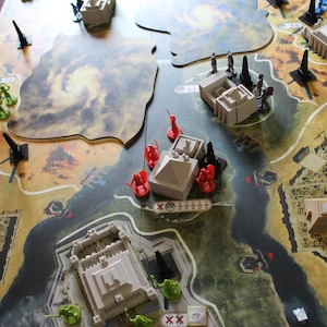 Pode incluir: Um jogo de tabuleiro com um tabuleiro em forma de mapa com edifícios em miniatura, pirâmides e figuras de várias cores. O tabuleiro tem um rio e um terreno semelhante ao deserto. Canhões e figuras em miniatura são colocados no tabuleiro.