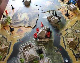 May include: A board game with a map-like game board featuring miniature buildings, pyramids, and figures in various colors. The game board has a river and desert-like terrain. Miniature cannons and figures are placed on the board, ready for play.