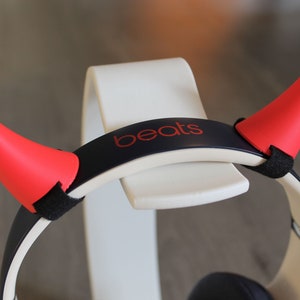 Puede incluir: Auriculares Beats negros con cuernos de diablo rojos unidos a la diadema. La diadema tiene la palabra "beats" impresa en rojo.