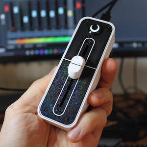 Puede incluir: Un mezclador de audio blanco y negro con un solo deslizador y un botón redondo con un símbolo de luna creciente. El mezclador tiene un fondo negro con una textura brillante.