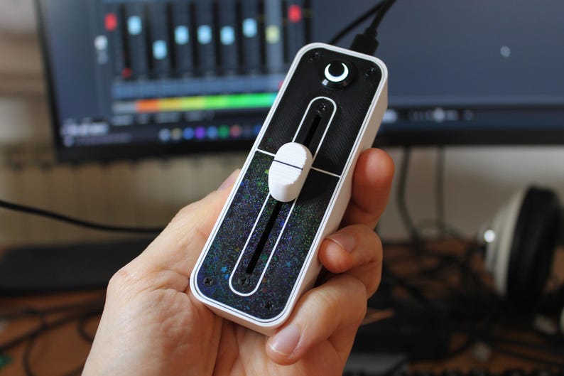 May include: A white and black audio mixer with a single slider and a round button with a crescent moon symbol. The mixer has a black background with a glittery texture.