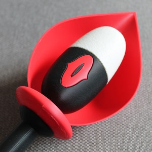 May include: A red and black bottle stopper with a white top. The stopper features a red lip design on a black background and is set in a red leaf-shaped holder. The handle is black.