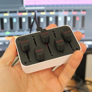 Op de afbeelding: Een klein, rechthoekig audiomixapparaat met vijf zwarte schuifregelaars en een witte basis. De schuifregelaars hebben rode accenten. Het apparaat wordt in een hand gehouden, met een computerscherm op de achtergrond.