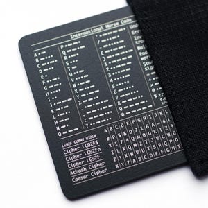 Peut inclure: Une carte rectangulaire en métal noir avec du texte et des diagrammes imprimés en blanc, incluant un tableau du code Morse international, des noms de chiffrements comme Atbash et César, et une grille de caractères alphanumériques, glissée dans une pochette en tissu noir.