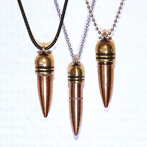 Peut inclure: Trois pendentifs en forme de balle en cuivre et laiton, chacun doté d'une pointe effilée et d'un capuchon arrondi avec des rainures horizontales. Un pendentif est suspendu à un cordon marron foncé, les deux autres à des chaînes à billes argentées.