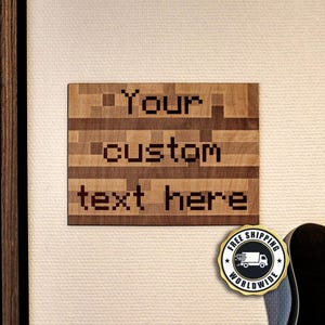 Könnte beinhalten: Ein rechteckiges Wand-Schild aus Holz mit einem pixeligen Blockmuster in verschiedenen Brauntönen. Der Text "Your custom text here" ist in einer dunklen, blockartigen Schrift im Pixel-Stil in die Mitte der Holzoberfläche eingraviert.