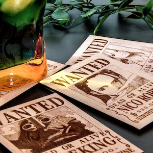 Puede incluir: Posavasos de madera con carteles de "Se Busca" con ilustraciones de personajes y texto. Los posavasos son de color marr&oacute;n claro y est&aacute;n dispuestos sobre una superficie oscura. Un vaso con l&iacute;quido verde y naranja est&aacute; en el fondo.
