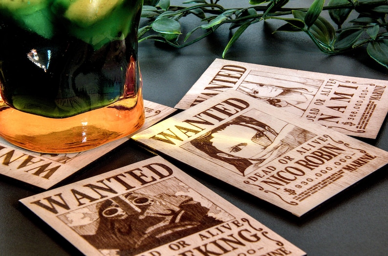 Puede incluir: Posavasos de madera con carteles de "Se Busca" con ilustraciones de personajes y texto. Los posavasos son de color marr&oacute;n claro y est&aacute;n dispuestos sobre una superficie oscura. Un vaso con l&iacute;quido verde y naranja est&aacute; en el fondo.