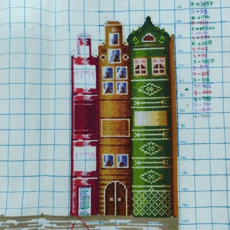 May include: A cross-stitch pattern of three buildings, each with a different colour scheme. The building on the left is red, the middle building is yellow, and the building on the right is green. Each building has a door and windows.