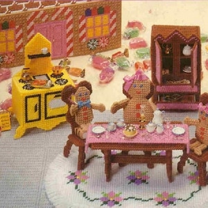 May include: A gingerbread house playset with a yellow stove, brown cabinet, and a table with four chairs. Four gingerbread people are sitting around the table. The set is made of plastic canvas and is perfect for a child's playroom.