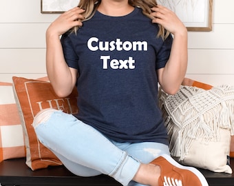 May include: A woman wearing a navy blue t-shirt with the text "Custom Text" in white. She is sitting on a bench with cushions and wearing light blue jeans and orange shoes.