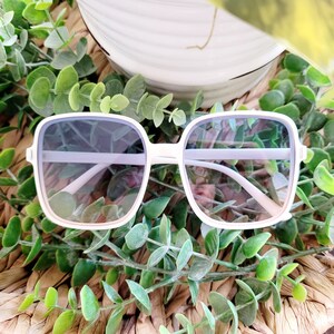May include: White square-shaped sunglasses with a light blue tint to the lenses. The sunglasses are sitting on a woven surface with green leafy plants.