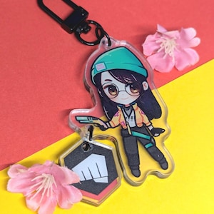 Könnte beinhalten: Ein Schlüsselanhänger mit einem Anime-Charakter in Grün und Gelb mit einem schwarzen und weißen Faustlogo. Der Charakter trägt eine grüne Mütze und hält eine Waffe. Der Schlüsselanhänger ist an einem schwarzen Metallring befestigt.