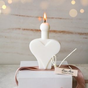 May include: A white, heart-shaped candle with a burning flame. The candle sits on a white box, adorned with a brown ribbon and a small tag that reads "Ersu Store Handmade with love."