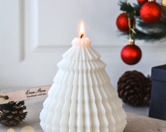 May include: A white, tree-shaped candle with a lit wick. The candle has a textured, pleated design. A small Christmas tree with red ornaments is in the background. A pine cone and a tag that says "Ensu Store Handmade with love" are also visible.