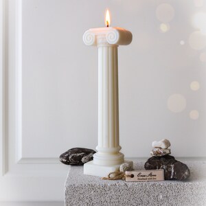 May include: A lit ivory pillar candle with a classical design. The candle has a fluted column and a decorative top. The candle is on a gray block with decorative stones and a tag that reads "Ersa Store, handmade with love."