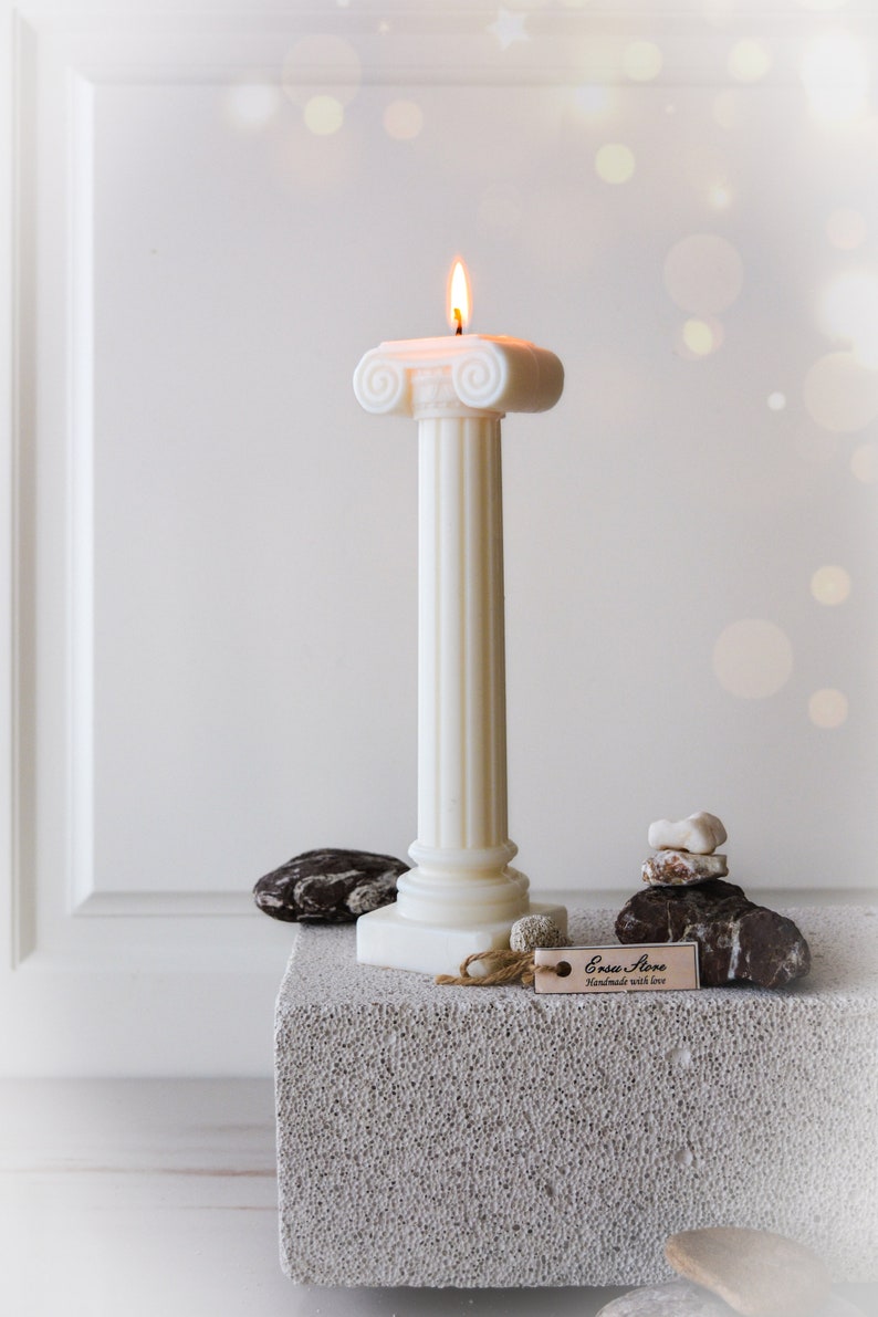 May include: A lit ivory pillar candle with a classical design. The candle has a fluted column and a decorative top. The candle is on a gray block with decorative stones and a tag that reads "Ersa Store, handmade with love."