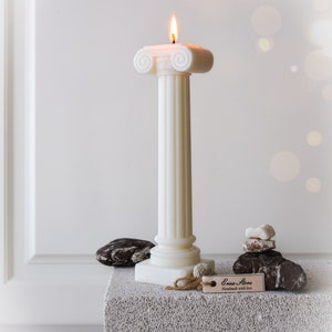 May include: A lit ivory pillar candle with a classical design, featuring a fluted column and a decorative top. The candle is placed on a textured gray surface, with decorative stones and a tag that reads "Ersa Store".