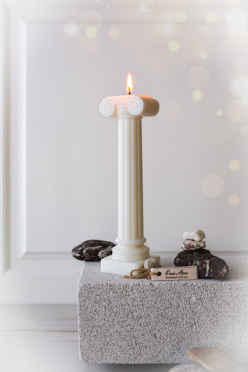 May include: A lit ivory pillar candle with a classical design, featuring a fluted column and a decorative top. The candle is placed on a textured gray surface, with decorative stones and a tag that reads "Ersa Store".