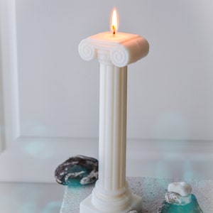 May include: A white pillar candle with a lit flame, designed in the style of a classical Greek column. The candle sits on a textured surface, with small stones and a tag nearby. The candle is handmade.
