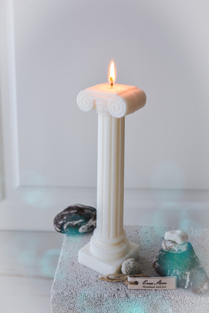 May include: A white pillar candle with a lit flame, designed in the style of a classical Greek column. The candle sits on a textured surface, with small stones and a tag nearby. The candle is handmade.