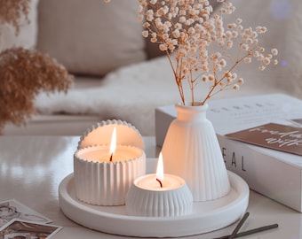 May include: A serene still life features white candles with lit flames, a small vase with delicate flowers, and a decorative tray. The scene is set on a white surface, with a soft, neutral background. The candles and vase are the focal point.