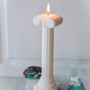 May include: A white pillar candle with a lit flame, designed in the style of a classical Greek column. The candle sits on a textured surface, with small stones and a tag nearby. The candle is handmade.