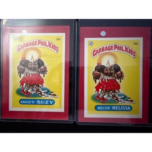 May include: Two Garbage Pail Kids trading cards, each in a protective sleeve. The cards feature a cartoon character with brown hair, a red dress, and oozing chocolate. One card reads "OOZY SUZY," the other "MELTIN' MELISSA."