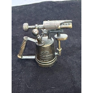 May include: An antique brass blowtorch with a ribbed handle, a nozzle, and a fuel tank. The blowtorch has a vintage aesthetic, with visible wear and tear. The text on the tank reads "RO.EY ESPAÑA".