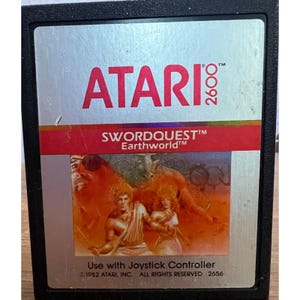 May include: A vintage Atari 2600 game cartridge for Swordquest: Earthworld. The cartridge is silver with red and black text and a colorful image of characters from the game.