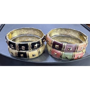 May include: Two sets of gold-toned metal bangles. One set features black, cream, and yellow enamel squares with gold studs. The other set has pink, purple, and green enamel squares with gold studs. Both sets have a textured gold band.