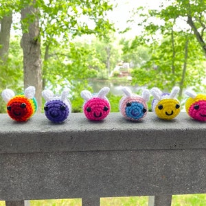 May include: Six crocheted bee plushies in a rainbow color scheme. The bees are lined up on a wooden railing with a green background.