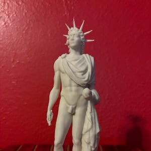 May include: White porcelain statue of a male figure with a crown of spikes on his head. The figure is standing with one hand on his hip and the other holding a drapery.
