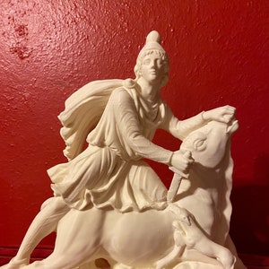 May include: A cream-coloured sculpture of a classical figure on horseback, wielding a sword. The sculpture has detailed carving and is set against a red background. The figure wears a helmet and cape.