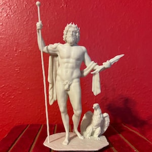 May include: A white matte figurine of a bearded deity wearing a crown and a draped cloth. The figure holds a tall staff in the right hand and a lightning bolt in the left, with an eagle standing at the feet on a hexagonal base.