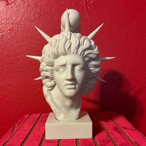May include: White marble bust sculpture of a woman with a crown of thorns on her head. The sculpture is on a white pedestal.