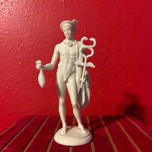May include: A white matte figurine of a standing figure wearing a winged hat and a draped cloth over one shoulder. The figure holds a small pouch in the right hand and a caduceus staff with two intertwined snakes and wings in the left hand.