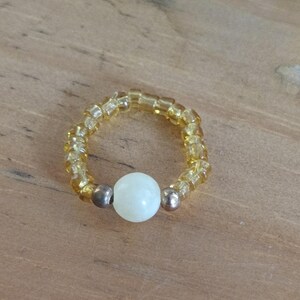 Peut inclure: Une bague perlée composée de petites perles ambrées et d'une perle centrale blanche et ronde. La bague a un design simple et élégant. La bague est présentée sur une surface en bois.