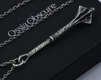 Puede incluir: Un colgante de metal plateado con forma de maza medieval, con un mango texturizado y una cabeza con bridas, suspendido de una cadena de eslabones a juego. La joya descansa sobre una caja negra con el texto Ossia Obscure impreso en blanco.