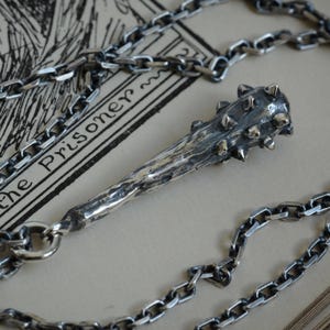 May include: A silver-toned metal pendant shaped like a spiked club or mace, featuring a textured, weathered surface and small protruding spikes at the top. The pendant is attached to a thick, interlocking silver-toned metal chain necklace.