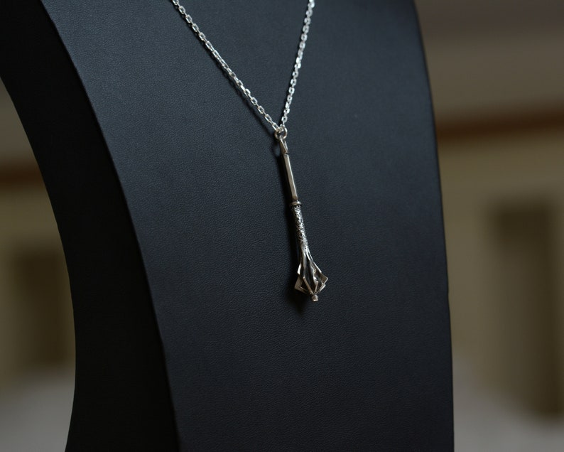 May include: A silver-toned pendant necklace featuring a mace-shaped charm with a textured handle and a flanged head. The pendant hangs from a delicate silver-colored chain, showcasing a metallic finish and intricate detailing on the weapon-inspired design.