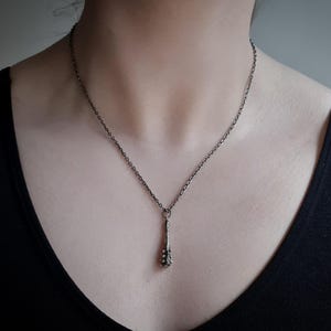 Puede incluir: Un collar de metal oscuro con un colgante delgado y texturizado en forma de peque&ntilde;a maza o garrote. El colgante cuelga de una cadena fina de metal oscuro a juego que descansa sobre el cuello, mostrando un acabado oxidado uniforme en toda la pieza.